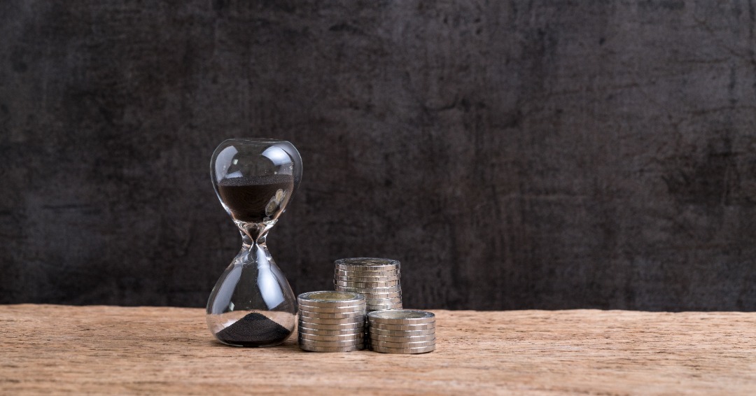 hour glass with black sand next to three stacks of coins to represent compound interest